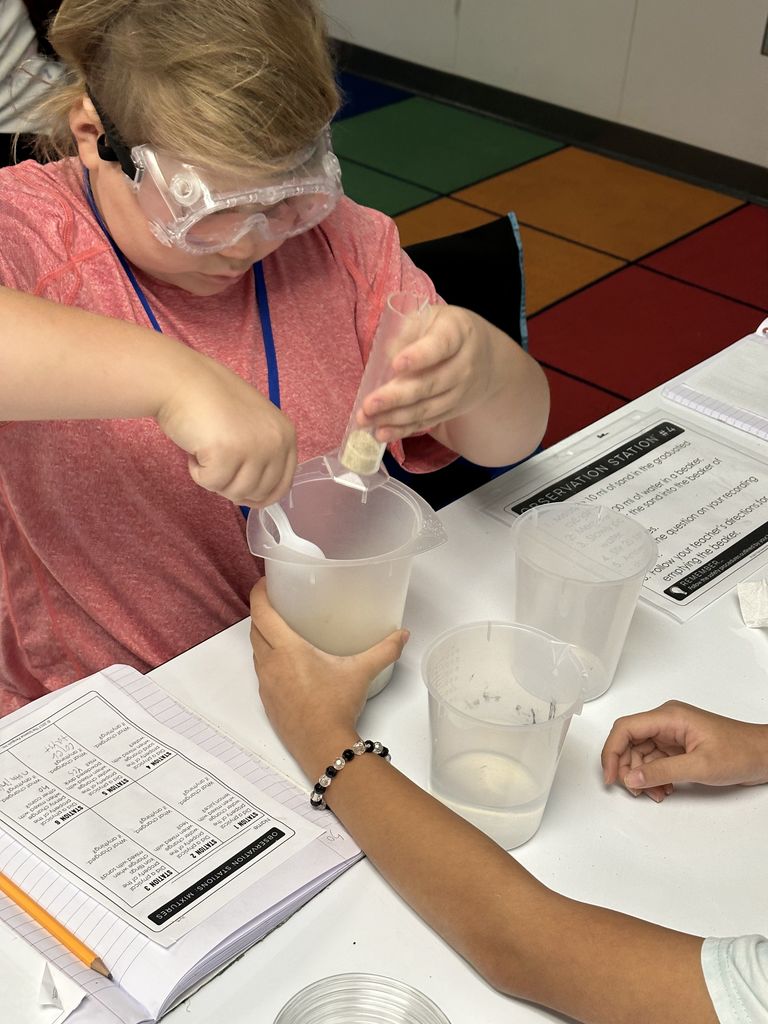 A boy wearing safety goggles  uses a plastic spoon and graduated cylinder to measure out sand. 